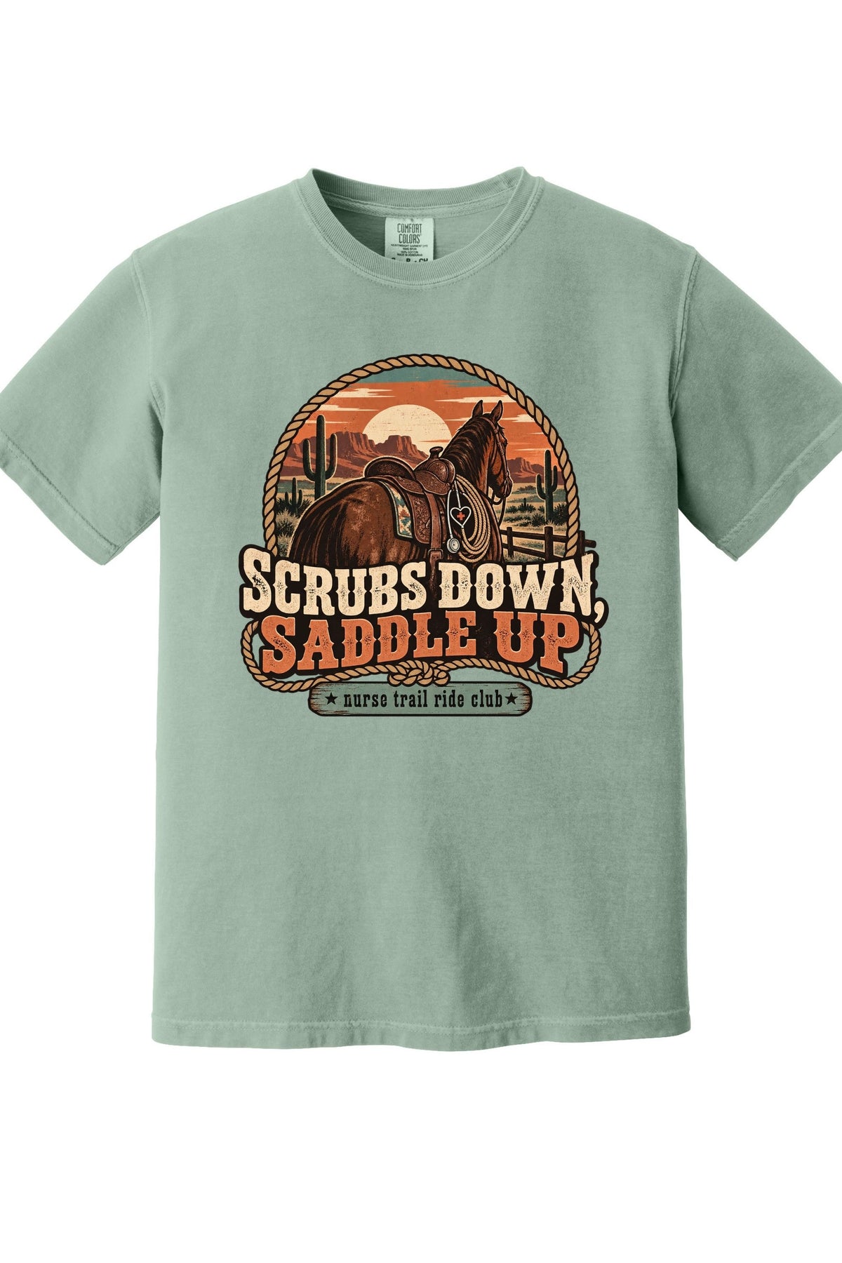 A sage green short-sleeve t-shirt with a retro western graphic of a horse and the text 'SCRUBS DOWN, SADDLE UP' and 'santa trail ride club'.