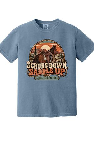 A light blue crewneck t-shirt featuring a western-themed graphic print with a horse, sunset, and the text 'SCRUBS DOWN, SADDLE UP' and 'survival ride club'.