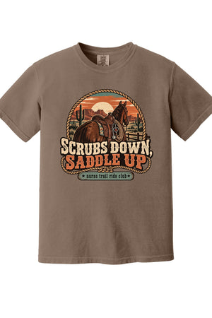 A brown short-sleeve crewneck t-shirt featuring a retro western graphic of a horse rider on a horse, with the text 'SCRUBS DOWN, SADDLE UP' and 'surf trail ride club' on a white background.