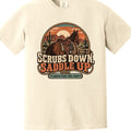 A cream-colored short-sleeve t-shirt with a large western-themed graphic on the chest featuring a horse, sunset, and the text 'SCRUBS DOWN SADDLE UP' and 'save trail ride club'.