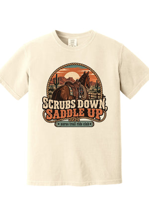 A cream-colored short-sleeve t-shirt with a large western-themed graphic on the chest featuring a horse, sunset, and the text 'SCRUBS DOWN SADDLE UP' and 'save trail ride club'.