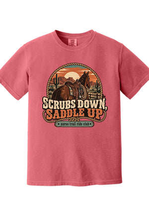 A pink short-sleeve crewneck t-shirt featuring a western-themed graphic print of a horse rider on a desert sunset with the text 'SCRUBS DOWN, SADDLE UP' and 'survival ride club'.